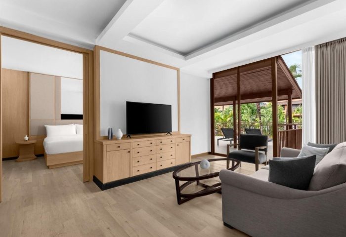 Courtyard Phuket, Patong Beach Resort suite interior. Living area with TV, couch, balcony view. Bedroom visible. Modern design, perfect for gay travel.
