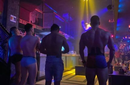 Gay strip bar in Puerto Vallarta. Muscular men in underwear and shorts at a gay party, with drinks and colorful lights. Gay travel nightlife.