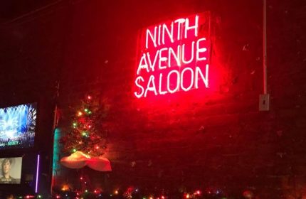Ninth Avenue Saloon, a gay bar in New York. Red neon sign on a brick wall, Christmas tree, and festive lights. Gay travel advice.