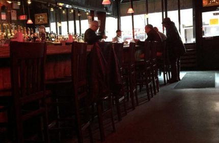 Interior of 9th Avenue Saloon, a gay bar in New York. People sit at the bar. A row of bar stools lines the counter in this NYC gay bar.