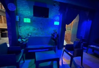 Adam Sauna Roma: Lounge area with blue lighting, chairs, tables, and a TV. A relaxing space for gay men's travel in Rome.