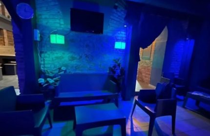 Adam Sauna Roma: Lounge area with blue lighting, chairs, tables, and a TV. A relaxing space for gay men's travel in Rome.