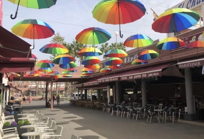 Adonis Bar, a gay bar in Gran Canaria, features rainbow umbrellas overhead. Tables and chairs line the street outside the bar.