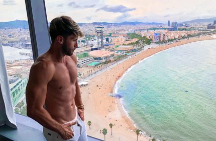 Gay-friendly W Barcelona hotel view. Fit man looks out over Barceloneta Beach and city skyline from adults-only hotel. Luxury gay travel.