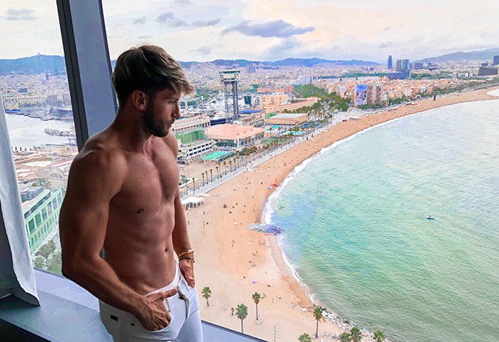 Gay-friendly W Barcelona hotel view. Fit man looks out over Barceloneta Beach and city skyline from adults-only hotel. Luxury gay travel.