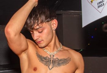 Shirtless man at Afterline Milan gay bar. He has a cross tattoo with wings, gold chain, and earring. Gay travel advice.
