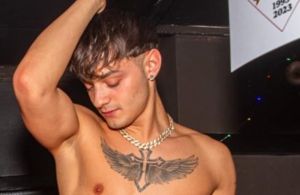 Shirtless man at Afterline Milan gay bar. He has a cross tattoo with wings, gold chain, and earring. Gay travel advice.