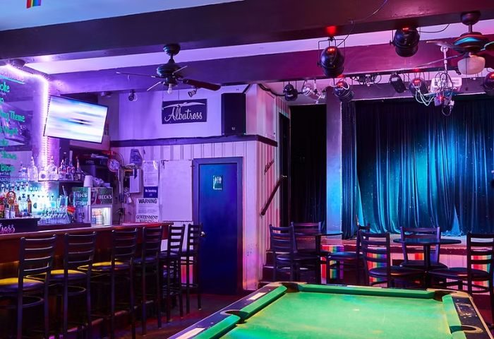 Interior of Albatross Bar in New York. A gay bar with pool table, stage, bar with stools, and a TV. Purple and blue lighting. Gay travel.