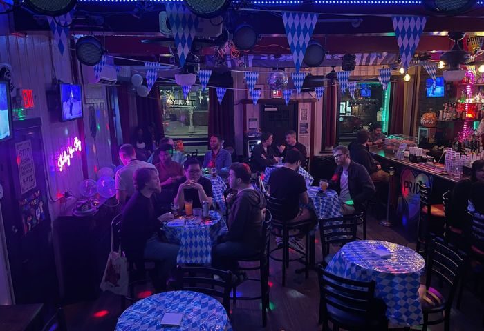 Inside Albatross Bar, New York. Gay bar scene with patrons at tables, bar with Pride sign, blue & white decor. Gay travel advice.