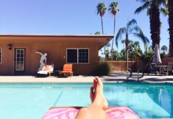 All-Worlds Resort gay rooms in Palm Springs. Person relaxing poolside at gay resort. Palm trees and blue sky in background. Vacation.