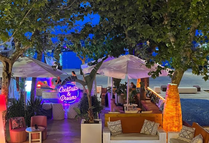 Angelo's Gay Bar in Ibiza at night. Outdoor seating area with Cocktails & Dreams neon sign, trees, and waterfront views. Gay travel.