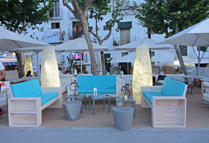 Angelo's Gay Bar in Ibiza. Outdoor seating area with blue cushions, hookahs, and white umbrellas. Gay travel destination, nightlife.