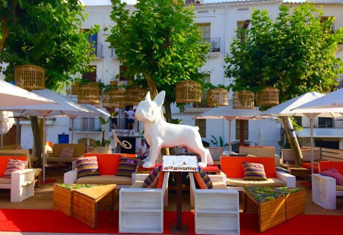 Angelo's Gay Bar in Ibiza. Outdoor seating area with white dog statue, colorful cushions, and birdcage decor. Gay travel destination.
