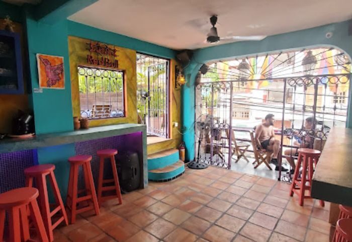 Anonimo Gay Bar in Puerto Vallarta, Mexico. Colorful bar interior with stools, open patio, and two men sitting outside. Gay travel.
