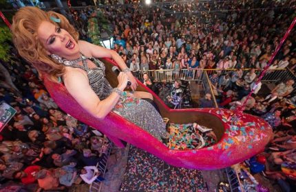 Aqua Bar Key West gay party: Drag queen in a pink shoe above a crowd. Nightclub scene with confetti. Gay travel destination.