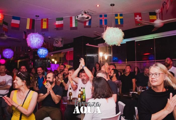 Aqua Club gay cruise club in Torremolinos. Cheering crowd at Terrazza Aqua, a popular gay bar. Flags and festive lights decorate the space.