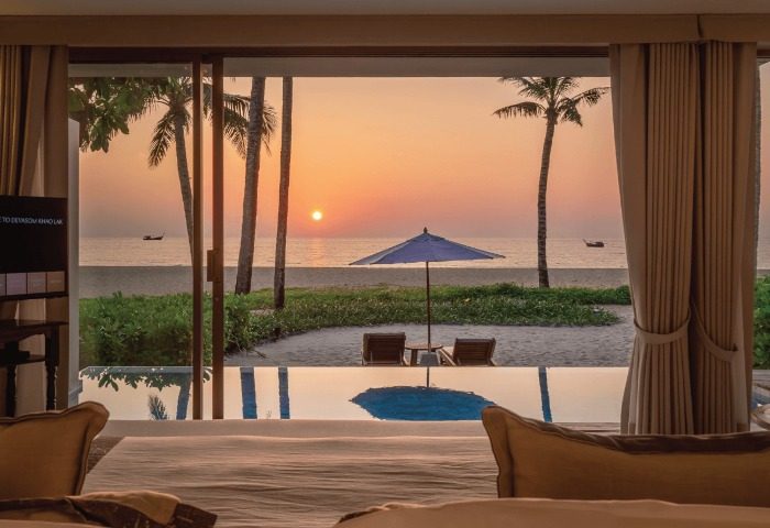 Devasom Khao Lak Beach Resort view. Sunset on beach with palm trees, umbrella, chairs. Villas' pool reflects the sky. Gay travel advice.