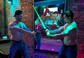 Shirtless men duel with lightsabers at Atmosphere, a gay live music bar in Andersonville, Chicago. Brick wall, mirror, and neon lights.