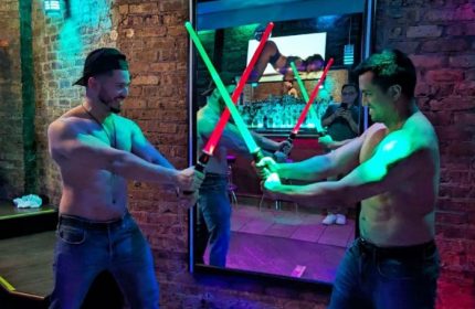 Shirtless men duel with lightsabers at Atmosphere, a gay live music bar in Andersonville, Chicago. Brick wall, mirror, and neon lights.