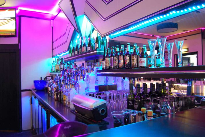 Bar at BJ's White Swan gay club. Shelves stocked with liquor bottles, glasses, and vibrant pink and blue neon lighting.