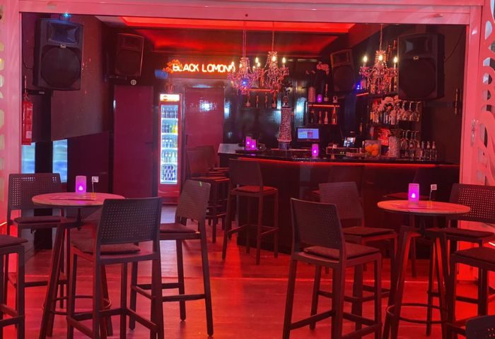 Interior of Black Lomond Bar, a gay bar in Gran Canaria. Red lighting, bar stools, tables, and a neon sign create a vibrant atmosphere.