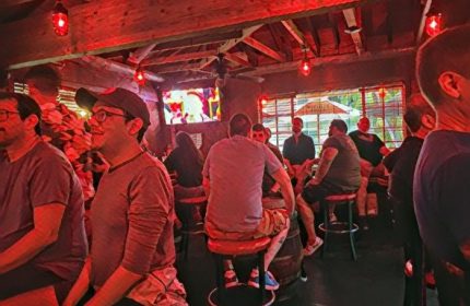 Inside Bacchus Waikiki gay bar in Honolulu. Men at bar stools, red lighting, and a TV screen. Popular gay party spot.