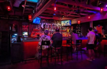 Bacchus Waikiki gay bar in Honolulu, a party spot. People at the bar, red lighting, TVs, and a lively atmosphere. Travel advice for gay men.