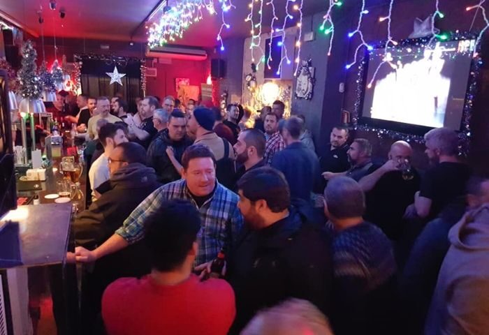 Crowded Bacon Bear Bar, a gay bar in Barcelona. Many men are standing, drinking, and socializing. Festive lights decorate the bar.