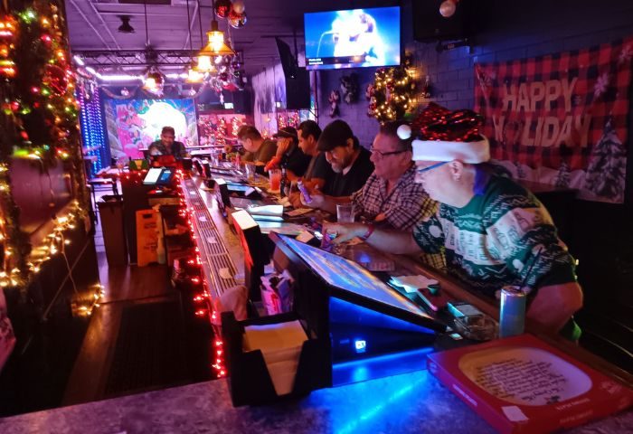 Holiday cheer at Badlands Las Vegas, a gay bar. Men are seated at the bar, decorated with lights. A TV screen shows a singer.