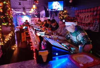 Holiday cheer at Badlands Las Vegas, a gay bar. Men are seated at the bar, decorated with lights. A TV screen shows a singer.