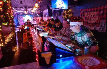 Holiday cheer at Badlands Las Vegas, a gay bar. Men are seated at the bar, decorated with lights. A TV screen shows a singer.