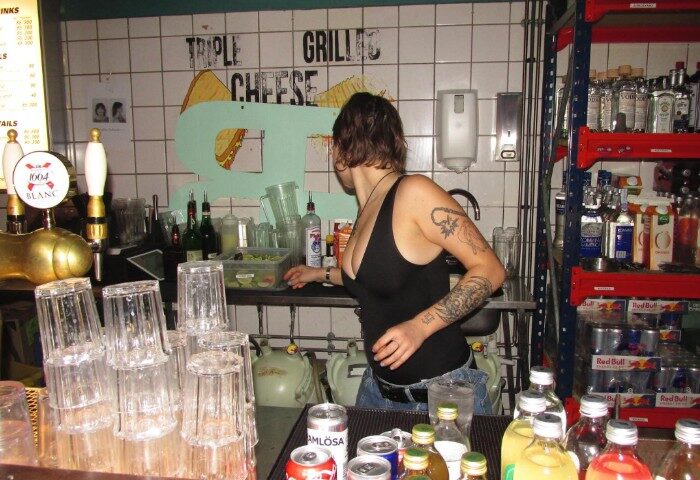 Baggen Copenhagen bar scene: Bartender with tattoos in a casual gay bar, drinks, glasses, and Triple Grilled Cheese sign.