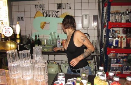 Baggen Copenhagen bar scene: Bartender with tattoos in a casual gay bar, drinks, glasses, and Triple Grilled Cheese sign.
