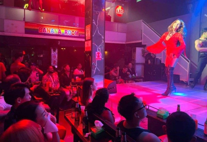 Banana Room Club, a gay bar in Bangkok, Thailand. Drag queen performing on stage for a seated audience. Nightlife scene, red lighting.