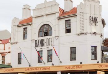 Bank Hotel, Newtown: White building with BANK HOTEL signage. Flags fly below, advertising live music, garden bar, and public bar. Gay travel.