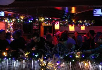 Montreal's Bar Aigle Noir gay bar party. Festive drinks, garland, and lights adorn the bar. People gather to celebrate. Gay travel.