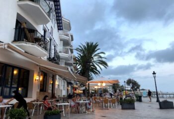 Outdoor seating at a gay bar in Sitges, Spain. People at tables, palm trees, and white buildings create a relaxed, vacation atmosphere.