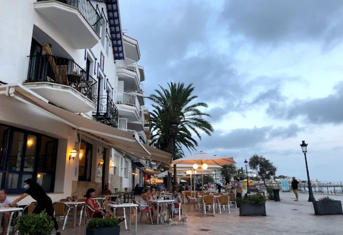 Outdoor seating at a gay bar in Sitges, Spain. People at tables, palm trees, and white buildings create a relaxed, vacation atmosphere.