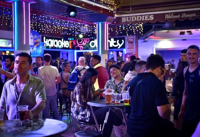 Nightlife at Bar Divinity, a gay bar in Gran Canaria. Crowded scene with men drinking, neon lights, and karaoke. Gay travel destination.