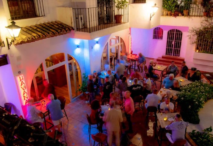 Gay bar in Torremolinos: Havana Bar's outdoor patio at night. People sit at tables, illuminated by colorful lights. Gay travel destination.