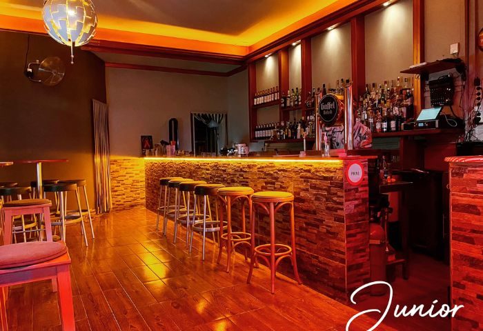 Interior of Junior Gay Bar in Gran Canaria. Bar stools line the textured bar front with a lit bar top. Bottles displayed on shelves.
