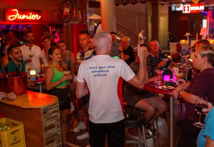 Inside Junior Gay Bar in Gran Canaria, a vibrant scene unfolds with patrons enjoying drinks at tables and the bar. A man with a t-shirt stands.