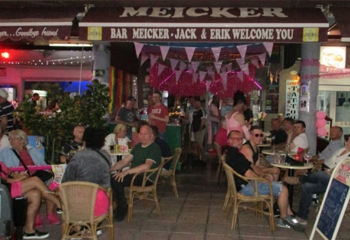 Exterior of Bar Meicker, a gay bar in Gran Canaria. Patrons sit at tables outside. Pink decorations adorn the entrance. Nightlife scene.