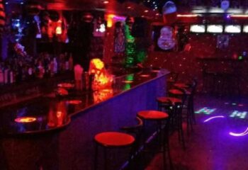 Inside the Mercury gay bar in Benidorm. Red bar stools line the counter, with colorful lights creating a party atmosphere.