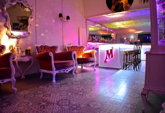 Interior of Bar Monalisa, a gay bar in Ibiza, with ornate chairs, a white bar with an M logo, and colorful lighting. Gay travel.