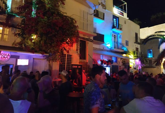 Night view of Bar Monalisa, a popular gay bar in Ibiza. Crowd of people outside, colorful lights, lively atmosphere. Gay travel destination.