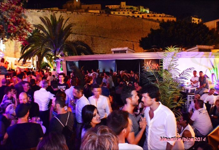 Night scene at Bar Mona Lisa, a gay bar in Ibiza. Crowd of people socializing, with the old town in the background. Gay travel advice.