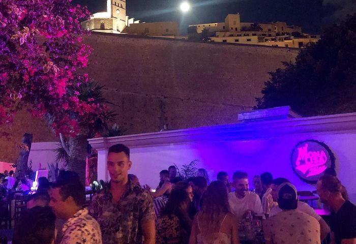 Night view of Bar Mona Lisa, a gay bar in Ibiza. Crowd of people, pink flowers, city view, and full moon. Gay travel destination.
