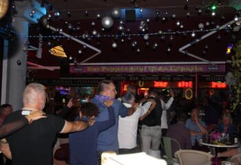 Gay bar scene at Bar Peppermint Gay Bar in Gran Canaria. Men with arms around each other, party lights, bar signs, and outdoor seating.