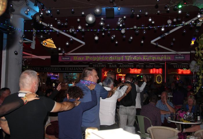 Gay bar scene at Bar Peppermint Gay Bar in Gran Canaria. Men with arms around each other, party lights, bar signs, and outdoor seating.